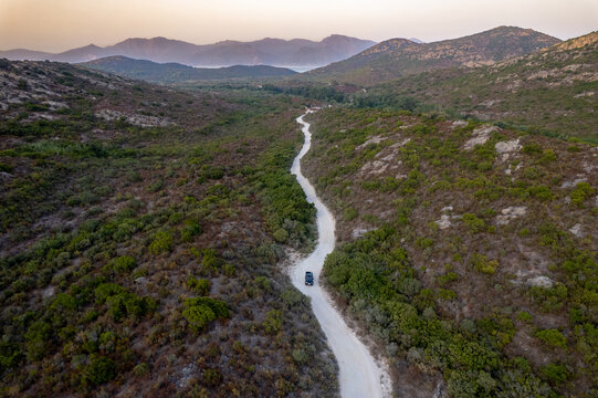 Aerial view of a winding dirt road cutting through the dense Mediterranean maquis leading towards the coast near Plage de Saleccia, with a vehicle driving along the path, Corsica, France.