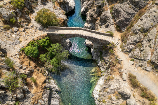 Aerial view of the historic Genoese bridge over the clear blue river in the rocky gorge of Asco, Corsica, France.