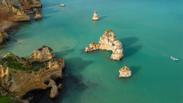 Aerial view of the limestone rock formations and sea stacks at Ponta da Piedade surrounded by turquoise water with a small boat cruising near the coast in Lagos, Faro District, Portugal.