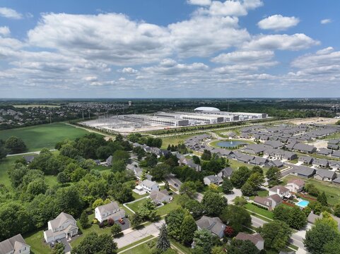 Aerial view of a large data center facility next to a suburban residential neighborhood with houses, green trees, and a small pond under a blue sky in the United States.