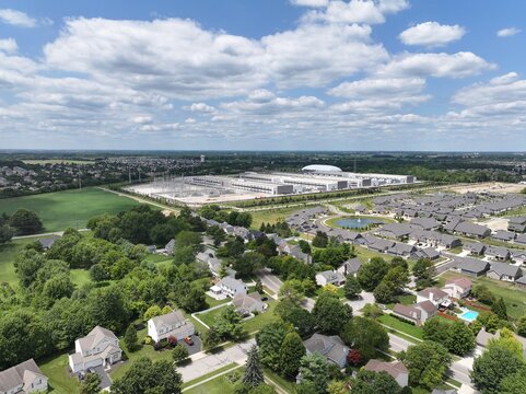 Aerial view of a large data center facility situated in a suburban landscape with residential houses, green trees, and a blue sky with clouds in the United States.