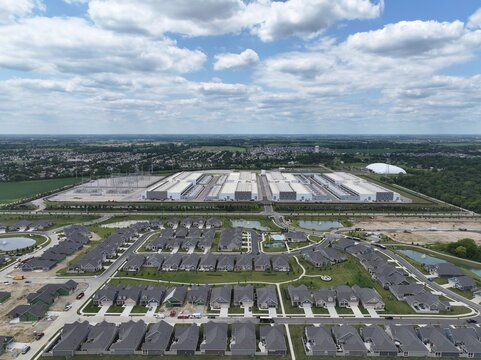 Aerial view of a large data center complex situated next to a modern suburban residential neighborhood with numerous houses, green spaces, and roads in the United States.