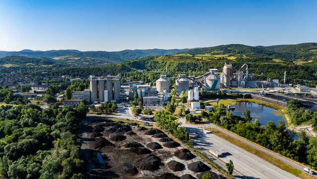 Aerial view of the Holcim Serbia cement factory with industrial silos, processing structures, and storage piles set against green hills and a pond in Beocin, Vojvodina, Serbia.