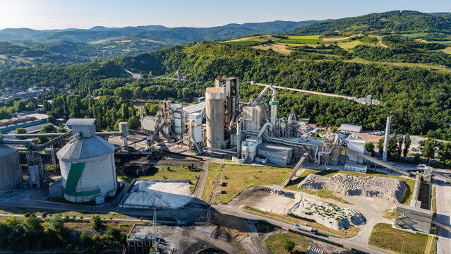 Aerial view of the Holcim Srbija cement factory featuring large industrial silos, processing structures, and storage areas surrounded by lush green hills in Beocin, Vojvodina, Serbia.