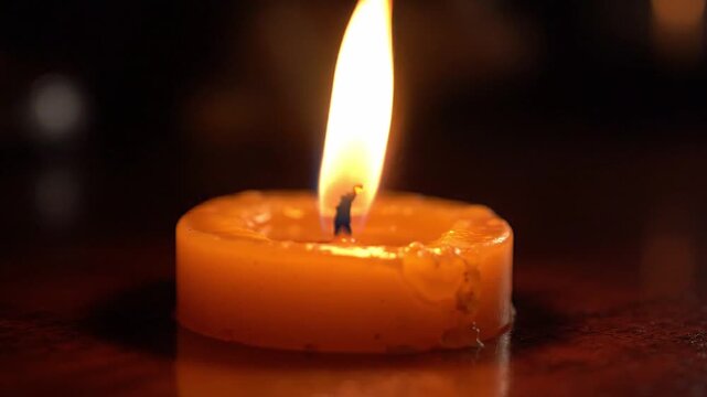 Candle flame flickers steadily in orange wax holder, showcasing gradual melting and wax pooling on wooden table surface in a dimly lit environment