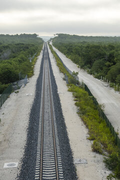 View of the Tren Maya railway tracks stretching through the dense jungle landscape with gravel ballast and adjacent dirt road in Calakmul, Campeche, Mexico.