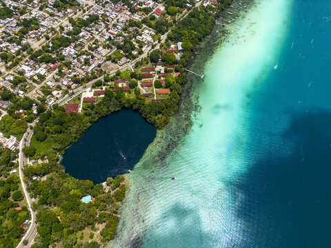 Aerial view of Cenote Negro with deep blue waters contrasting against the turquoise shallows of Lake Bacalar, surrounded by lush vegetation and town buildings, Bacalar, Quintana Roo, Mexico.