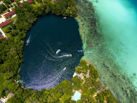 Aerial view of Cenote Negro with deep blue waters contrasting against the shallow turquoise lagoon, surrounded by lush tropical vegetation and boats in Bacalar, Quintana Roo, Mexico.