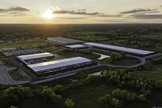 Aerial view of a modern data center complex with large flat-roofed buildings, surrounding green fields, and a power substation during a golden sunset in Hoffman Estates, Illinois, United States.