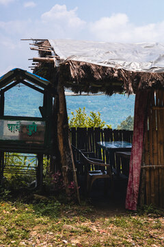 View of a rustic roadside stall with a thatched roof, plastic tarp, and bamboo walls featuring simple plastic furniture overlooking the lush green hills of Pokhara, Gandaki Province, Nepal.