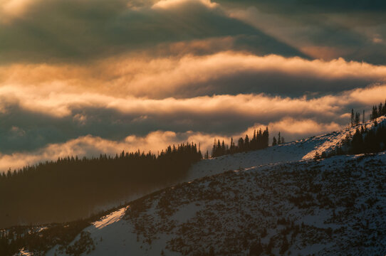 Aerial view of the snow-covered slopes of Dumbier in the Low Tatras mountains, featuring pine forests and dramatic golden hour clouds over Liptovsky Jan, Zilina Region, Slovakia.