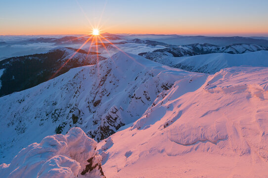 View of a sunrise over the snow-covered peaks of the Low Tatras mountains with a sunburst and clouds in the valley in Liptovsky Jan, Zilina Region, Slovakia.