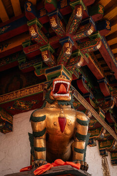 View of a golden snow lion statue with an open mouth and orange cloth, set against ornate, colorful painted wooden ceiling beams in a Tibetan monastery in Lhasa, Tibet, China.