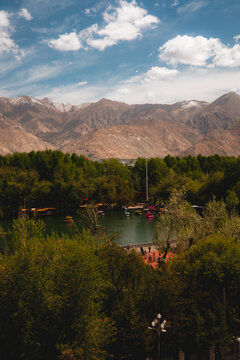 View of a tranquil lake in a park surrounded by lush trees and rugged mountains under a blue sky with scattered clouds in Lhasa, Tibet, China.