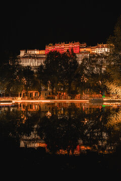 View of the illuminated Potala Palace at night with its reflection shimmering in the foreground water, surrounded by dark trees in Lhasa, Tibet, China.