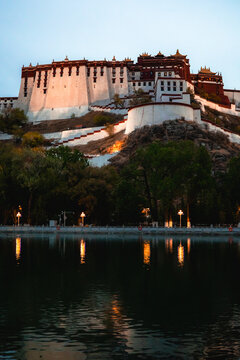 View of Potala Palace illuminated at dusk with its reflection shimmering in the water and surrounding trees in Lhasa, Tibet, China.