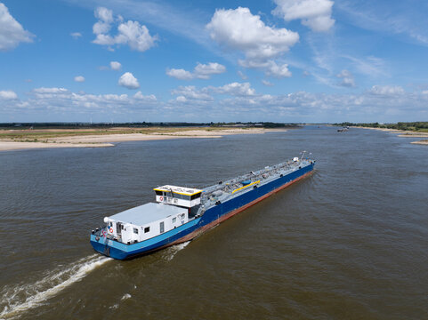 Aerial view of a blue and white inland tanker ship navigating a wide river under a bright blue sky with scattered clouds in the Netherlands.