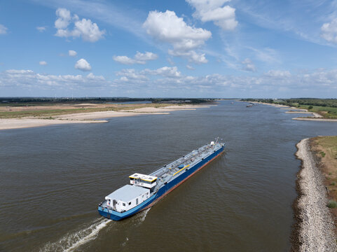 Aerial view of a large cargo barge navigating a wide river with grassy banks and a blue sky with clouds in the Netherlands.