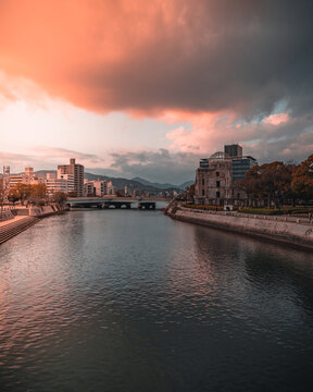 View of the Hiroshima Peace Memorial, also known as the Atomic Bomb Dome, along the Motoyasu River with urban buildings and a dramatic sunset sky in Hiroshima, Hiroshima Prefecture, Japan.