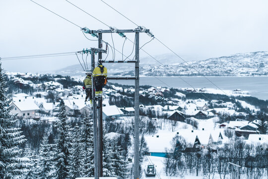 Tromso, Troms, Norway - 05 February 2020: View of two workers in high-visibility gear repairing power lines in a snowy landscape with houses and a fjord in the background.