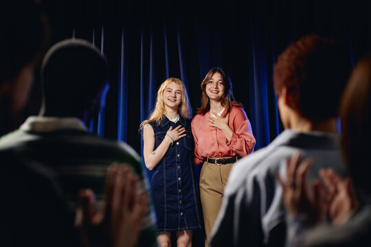 Portrait of Caucasian girls standing on stage smiling during student theater performance while audience applauds. Image useful for high school drama club and arts education marketing