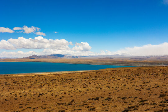View of Peiku Co, a high-altitude alpine lake surrounded by arid plains and distant snow-capped mountains under a clear blue sky in Shigatse, Tibet, China.