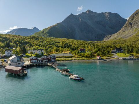 Aerial view of Koppangen Brygger with red wooden cabins on stilts over turquoise water, surrounded by lush forests and mountains in Lyngseidet, Troms, Norway.