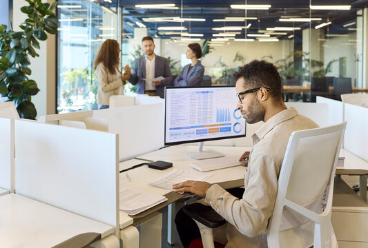 Analyst businessman manager in office reviewing data charts. He works at a computer with a spreadsheet as colleagues talk strategy nearby. Clear collaboration and finance analytics concept.