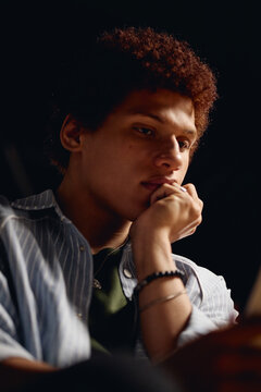 Young adult male student sitting in dark theater, holding chin in hand while reading script. Image is suitable for promoting youth arts programs, vertical
