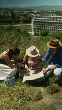 Three young tourists sit on a grassy hill, carefully studying a paper map. A group of friends are actively planning their next exploration during their vacation.