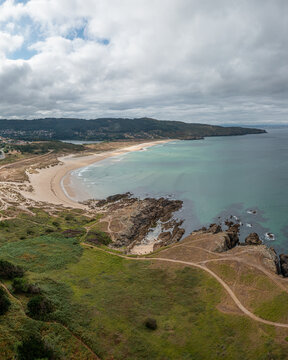 Aerial view of the sandy beach and rocky coastline of Valdovino with green hills and rolling waves under a cloudy sky in Valdovino, A Coruna, Spain.