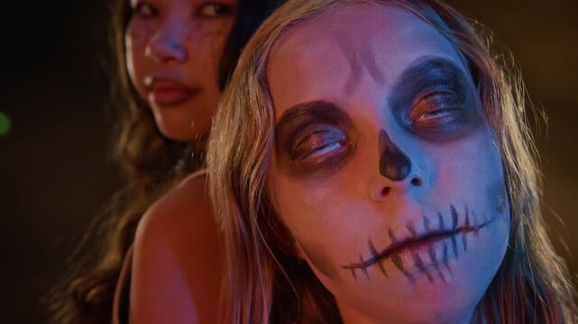 Multiethnic teenagers posing with Halloween makeup, Asian girl looking at camera while Caucasian girl with skull face paint tilting head down with eyes rolled. Ideal for holiday marketing campaigns
