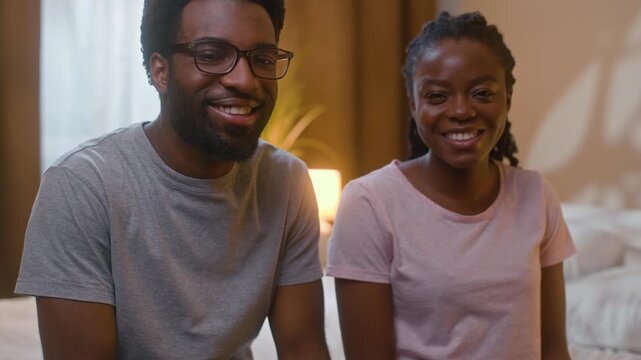 African American happy married couple smiling sitting on bed looking at camera wife husband loving family portrait ethnic spouses marriage relationship indoors home illuminated bedroom late evening