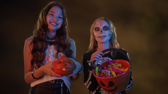 Portrait of Asian teenager and Caucasian teenager in skeleton makeup holding carved pumpkin and candy bucket, smiling at camera. Ideal for Halloween marketing and seasonal holiday promotions