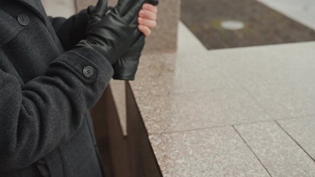 Man leaning on granite ledge, waiting with hands in pockets removed leather gloves, beanie and scarf, drizzly pavement and muted city backdrop, candid lifestyle broll capturing creator pause and quiet
