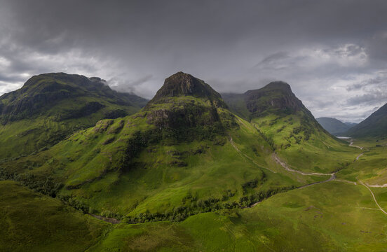 Aerial view of the Scottish Highlands featuring lush green mountain slopes, dramatic rocky peaks, and a winding stream under a moody, overcast sky, Scotland.