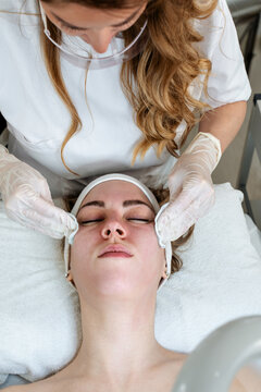 Professional beautician wearing face shield and gloves performs a deep cleansing facial treatment on a client in a spa clinic, focusing on skincare and hygiene.