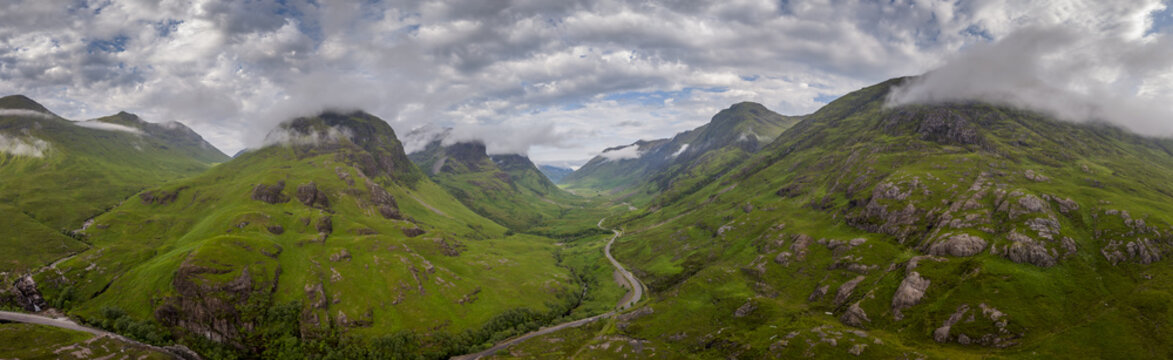 Aerial view of the Scottish Highlands featuring lush green mountains, a winding road through the valley, and rocky slopes under a cloudy sky in Scotland.