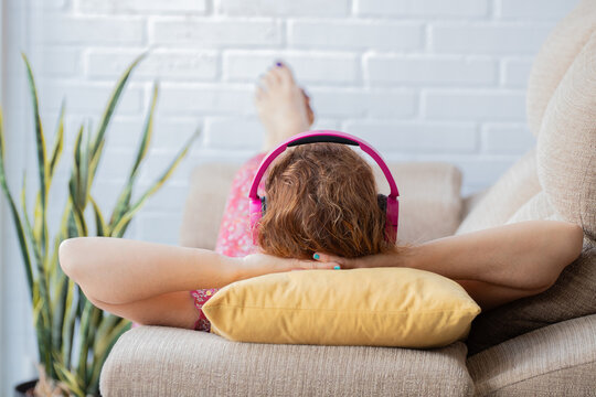 Young woman lying on a couch with pink headphones, hands behind her head, enjoying music in a bright living room