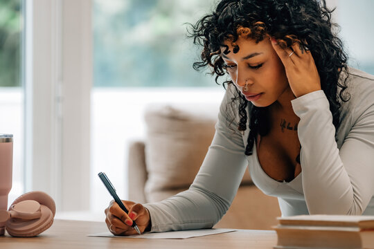 Focused young Black woman leaning over a desk, writing notes with pink headphones and tumbler nearby, studying or journaling at home