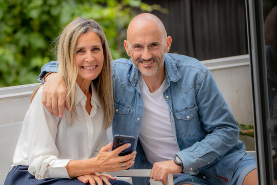 Happy caucasian couple in their 50s sharing a joyful moment outdoors, looking at a phone screen together, sitting in a sunny garden setting