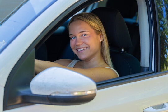 Happy young Caucasian woman smiling and driving a car, looking out the window with a cheerful, confident expression on a sunny day