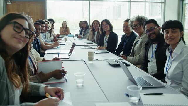 A diverse group of professionals seated around a conference table looking towards the camera, then happily applaud and smile, celebrating success or achievement during a meeting.