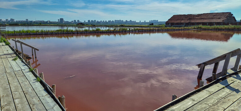 Wooden pier extending into a pink salt lake with a distant city skyline and abandoned building under a clear sky, near Burgas.