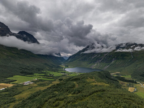 Aerial view of a lush green valley featuring a calm lake surrounded by steep mountains partially covered in low-hanging clouds under a dramatic overcast sky, Norway.