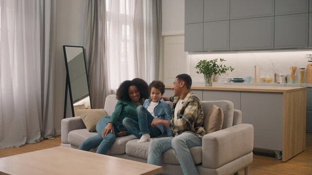 Happy excited African American family mom dad son jumping sofa in apartment relocation woman female little boy child kid father male man moving couch furniture fall celebrating buying new home indoor
