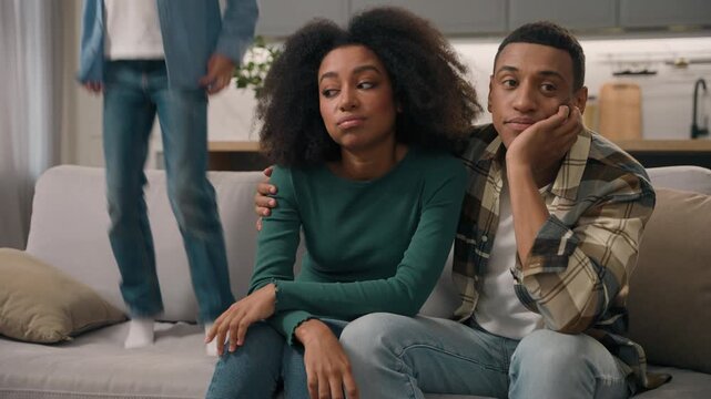 Sad African American family parents hugging on couch unhappy mother father and loud little boy child son jumping sofa home room desperate tired mom dad ignoring misbehavior active kid shouting problem