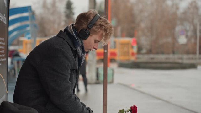 White creator gazing streetcar holding rose, wearing heavy coat and headphones profile shot captures hopeful expression as urban traffic and passersby blur in background, subtle narrative