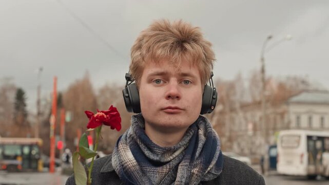 White creator in headphones with rose, playful street performer offering bloom then making rude gesture, autumn bus stop scene, scarfed artist with deadpan expression and city buses behind.