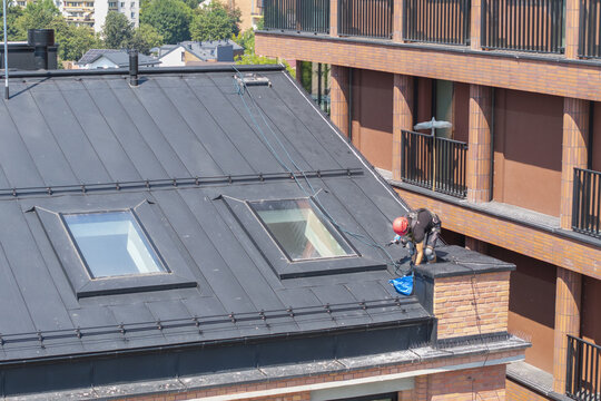 Aerial view of a worker in a red helmet and safety harness working on a dark metal roof with skylights next to a brick building in Krakow, Lesser Poland Voivode, Poland.